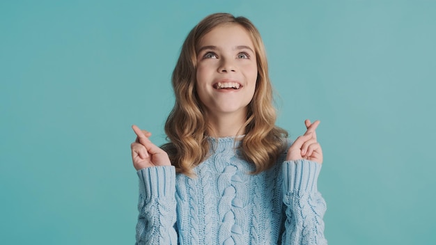 Una adolescente rubia muy emocionada que se ve feliz manteniendo los dedos cruzados, espero que sus sueños se hagan realidad sobre el fondo azul