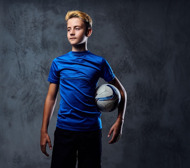 Foto gratuita adolescente rubio, jugador de fútbol vestido con un uniforme azul sostiene una pelota.