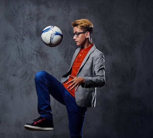 Un adolescente rubio vestido con jeans juega con una pelota de fútbol sobre un fondo gris.