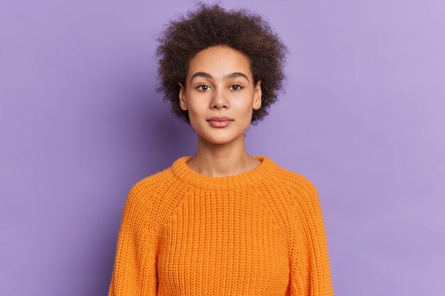 Una adolescente seria de piel oscura con cabello rizado y tupido se ve con confianza y tiene una expresión tranquila de belleza natural vestida con suéter de punto.