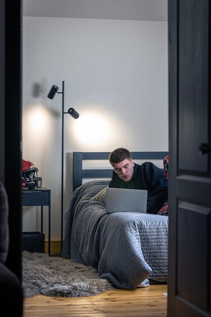 Un adolescente se sienta en una habitación en una cama y usa una computadora portátil