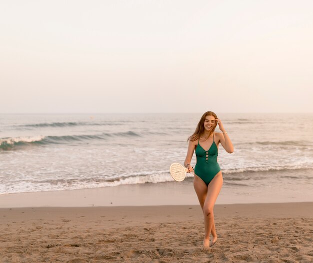 Adolescente sonriente en bikini verde sosteniendo la raqueta de pie cerca de la orilla del mar