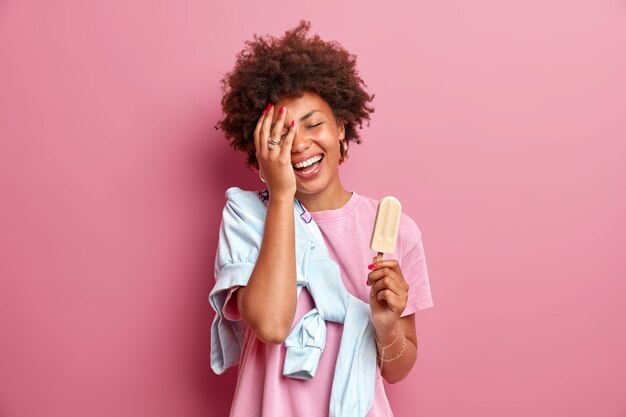 La adolescente sonriente hace que la palma de la cara se sienta feliz, usa una camiseta casual con un puente, disfruta del horario de verano, come un delicioso helado en un palo, tiene buen humor aislado sobre una pared rosa. Diversión y dulces