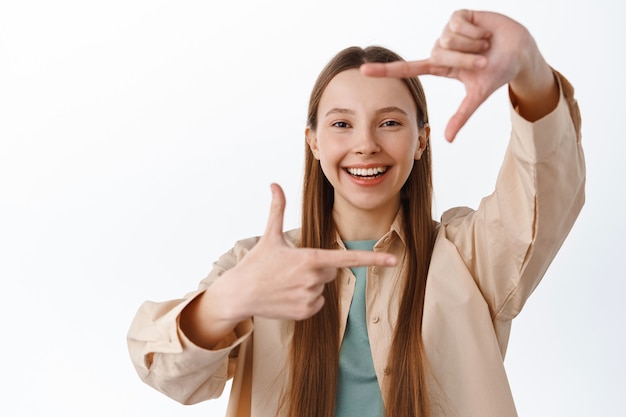 Adolescente sonriente mira a través de marcos de mano gesto de cámara, imaginando algo, mide el ángulo perfecto, de pie sobre una pared blanca.