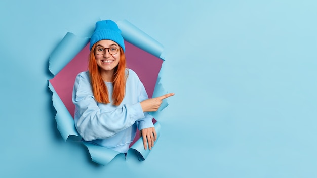 Adolescente sonriente con el pelo rojo da una recomendación, apuntando al espacio de la copia, rompe el agujero del papel azul