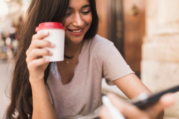 Adolescente sonriente que sostiene la taza de café disponible usando el teléfono móvil