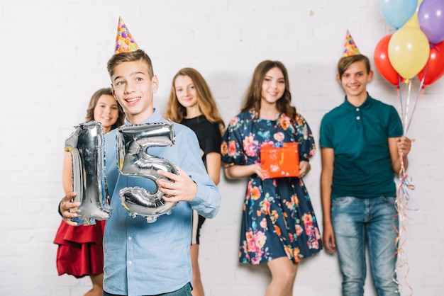 Un adolescente sosteniendo un número gris de globo de aluminio de 15, parado frente a sus amigos