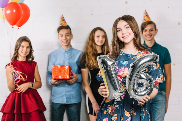 Una adolescente sosteniendo un número gris de globo de aluminio de 16, de pie frente a sus amigos