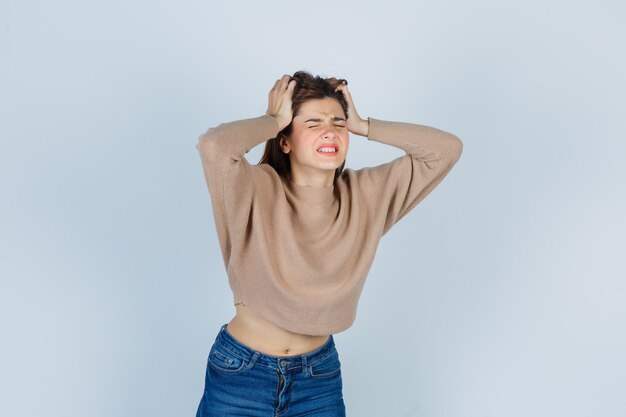 Adolescente en suéter, jeans agarrando la cabeza con las manos y mirando irritado, vista frontal.