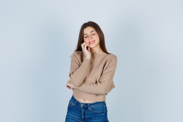 Adolescente en suéter, jeans inclinando la mejilla en la mano, cerrando los ojos y mirando soñadora, vista frontal.