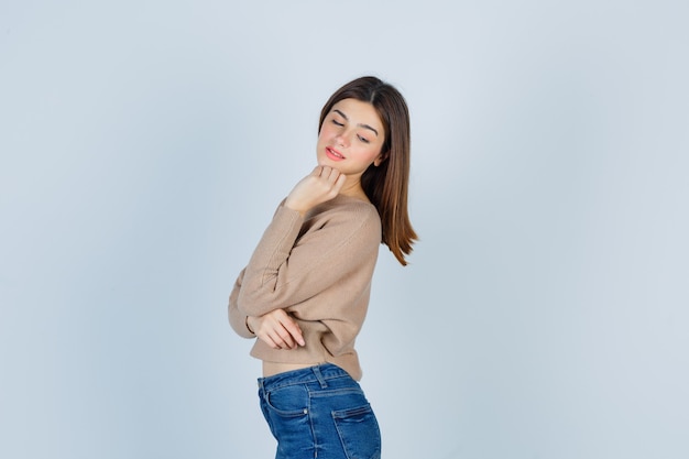 Adolescente en suéter, jeans manteniendo la mano en la barbilla, mirando hacia abajo y luciendo elegante.