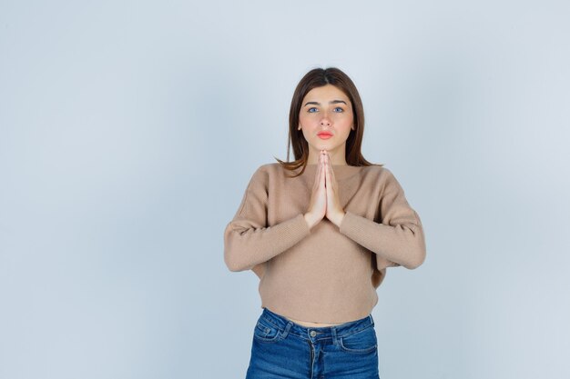 Adolescente en suéter, jeans mostrando las manos juntas en gesto de súplica y mirando esperanzado, vista frontal.