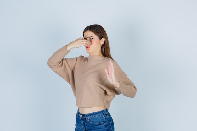 Adolescente en suéter, jeans oliendo algo desagradable, pellizcando la nariz, mostrando la señal de stop y mirando disgustado, vista frontal.