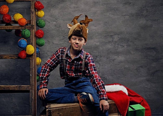 Adolescente vestido con una camisa a cuadros y un sombrero con cuerno de ciervo posando sobre un fondo decorado de Navidad.