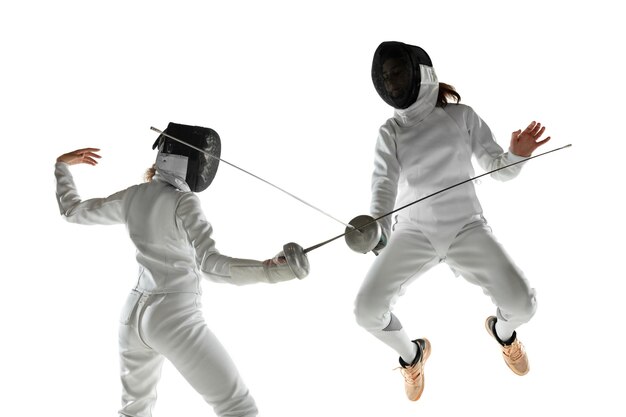 Las adolescentes en trajes de esgrima con espadas en las manos aisladas sobre fondo blanco de estudio. Modelos femeninos jóvenes practicando y entrenando en movimiento, acción. Copyspace.