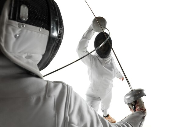 Las adolescentes en trajes de esgrima con espadas en las manos aisladas sobre fondo blanco de estudio. Modelos femeninos jóvenes practicando y entrenando en movimiento, acción. Copyspace.