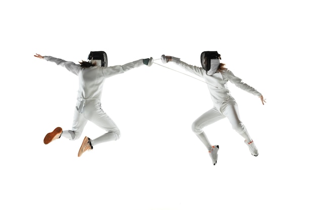 Las adolescentes en trajes de esgrima con espadas en las manos aisladas sobre fondo blanco de estudio. Modelos femeninos jóvenes practicando y entrenando en movimiento, acción. Copyspace.