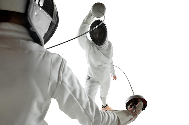 Las adolescentes en trajes de esgrima con espadas en las manos aisladas sobre fondo blanco de estudio. Modelos femeninos jóvenes practicando y entrenando en movimiento, acción. Copyspace.