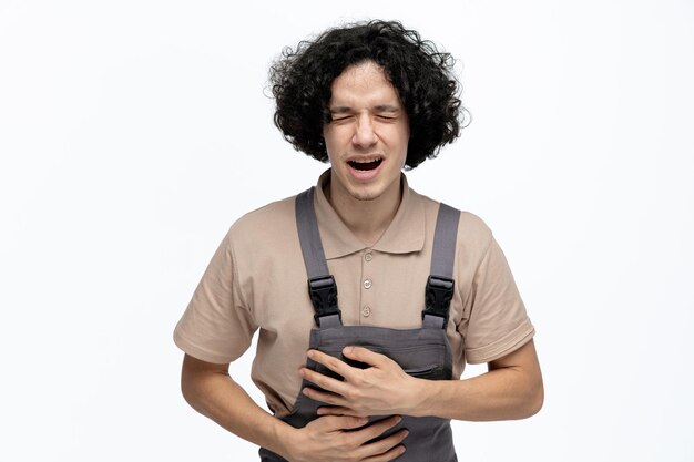 Adolorido joven trabajador de la construcción vistiendo uniforme manteniendo las manos en el vientre con los ojos cerrados aislado sobre fondo blanco.