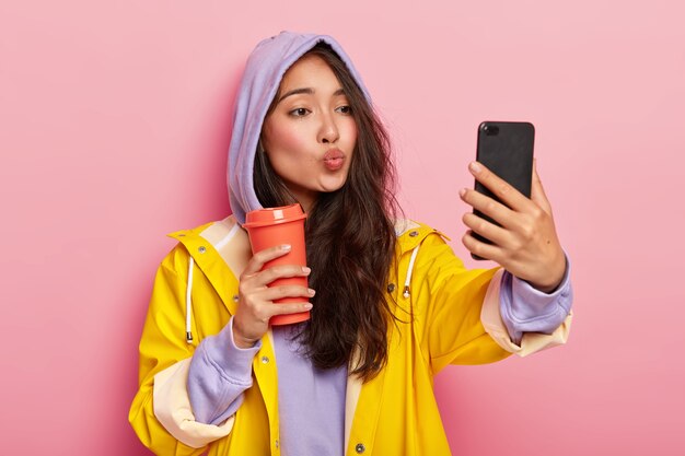 Adorable adolescente con apariencia asiática, mantiene los labios doblados, envía un beso a la cámara del teléfono celular, se toma una selfie, disfruta de la bebida, usa una sudadera, una capucha en la cabeza, un impermeable amarillo, camina después de la lluvia