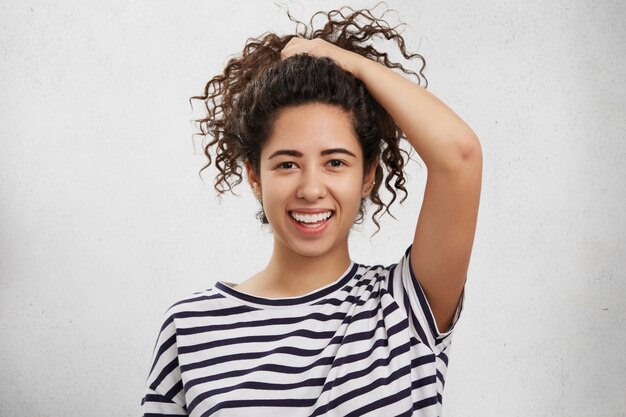 Adorable adolescente con cabello rizado y nítido, viste camisa a rayas, hace cola de caballo, anuncia el efecto agradable del nuevo champú