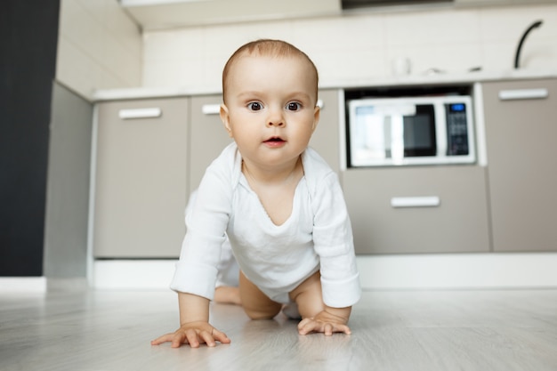 Adorable bebé gateando sobre el piso de la cocina con cara divertida