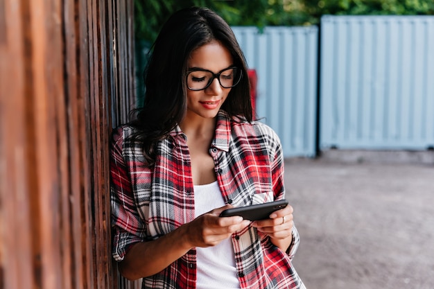 Foto gratuita adorable chica bronceada usando internet móvil con sonrisa. retrato al aire libre de alegre estudiante morena de pie en la calle con teléfono.