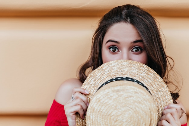 Adorable chica de cabello oscuro posando delante de la pared con expresión de sorpresa. Tiro al aire libre de la dama morena de buen humor que se esconde detrás del sombrero de verano.