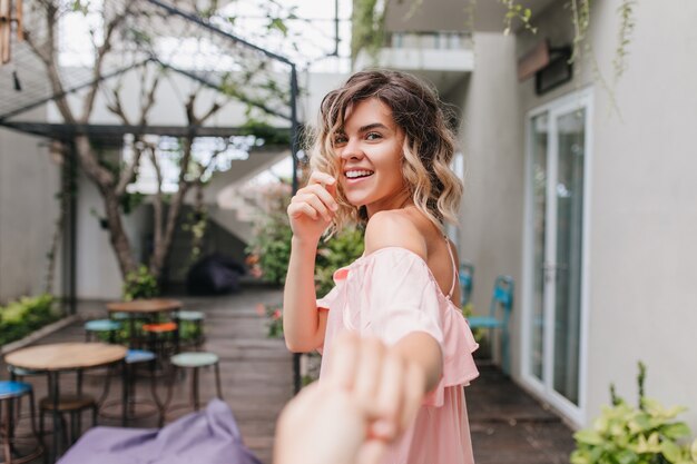 Adorable chica caucásica en traje rosa de moda disfrutando de la mañana del fin de semana en la cafetería de la calle. Tiro al aire libre de la dama europea rizada que expresa felicidad en vacaciones.