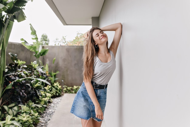 Adorable chica en falda de mezclilla relajante cerca de plantas. Retrato al aire libre de una mujer europea sonriendo por la mañana mientras está de pie junto a la pared blanca.