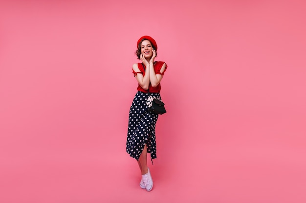 Adorable chica francesa en zapatos blancos posando con una sonrisa sincera. Toma interior de cuerpo entero de mujer de pelo corto despreocupada en boina roja.