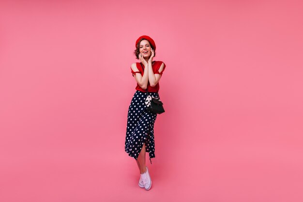 Adorable chica francesa en zapatos blancos posando con una sonrisa sincera. Toma interior de cuerpo entero de mujer de pelo corto despreocupada en boina roja.