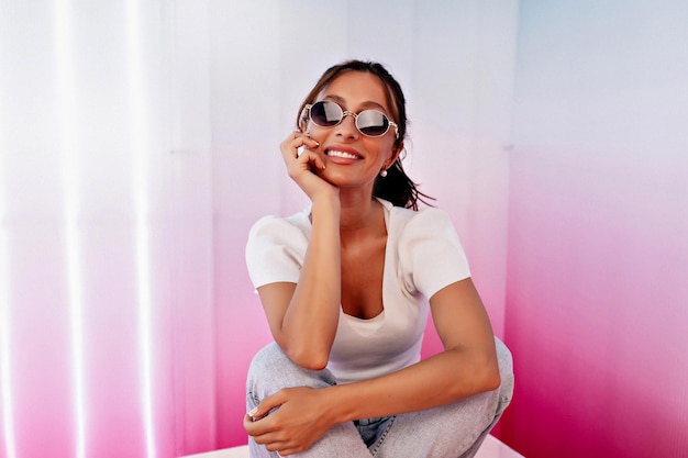 Adorable chica guapa con el pelo oscuro con gafas de sol posando en la cámara con una sonrisa feliz y amistosa mientras se sienta en un fondo rosa