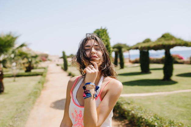 Adorable chica morena encantadora en camiseta blanca sin mangas y accesorios de moda posando con los ojos cerrados con árboles exóticos. Retrato de encantadora joven bronceada pasar tiempo afuera en la mañana