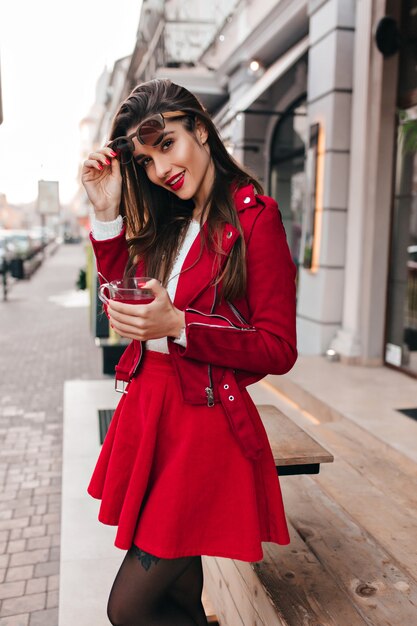 Adorable chica morena en falda roja disfrutando de una sesión de fotos al aire libre