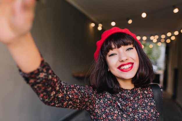 Adorable chica morena francesa con maquillaje elegante y peinado corto divirtiéndose con la cámara sobre fondo borroso. Bastante morena joven en ropa vintage haciendo selfie y sonriendo felizmente