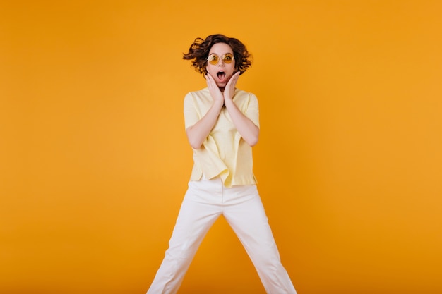 Adorable chica morena lleva gafas de sol naranjas saltando con expresión de asombro. Foto de una elegante dama blanca con una camiseta amarilla de gran tamaño.