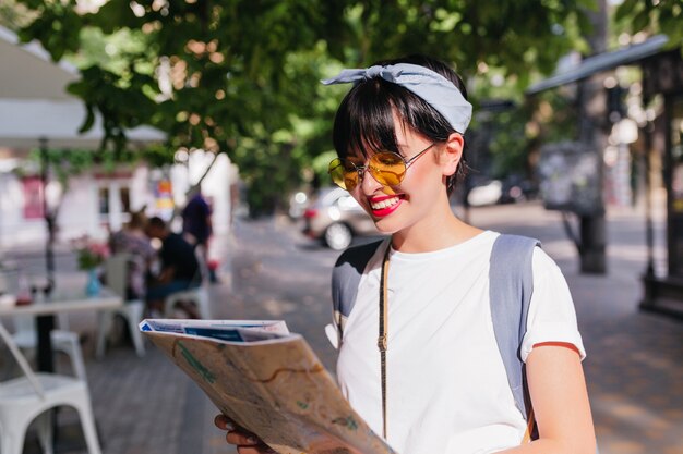 Adorable chica morena con sonrisa de hollywood mirando el mapa de la ciudad en busca de destino de pie en medio de la calle