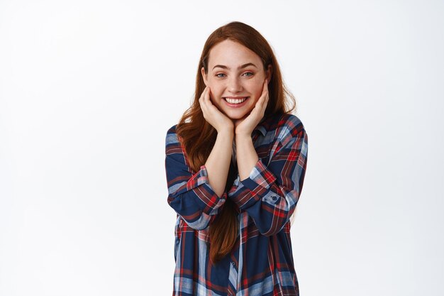 Adorable chica pelirroja suspirando y luciendo divertida, viendo algo interesante y emocionante, sonriendo divertida, parada en camisa a cuadros contra fondo blanco