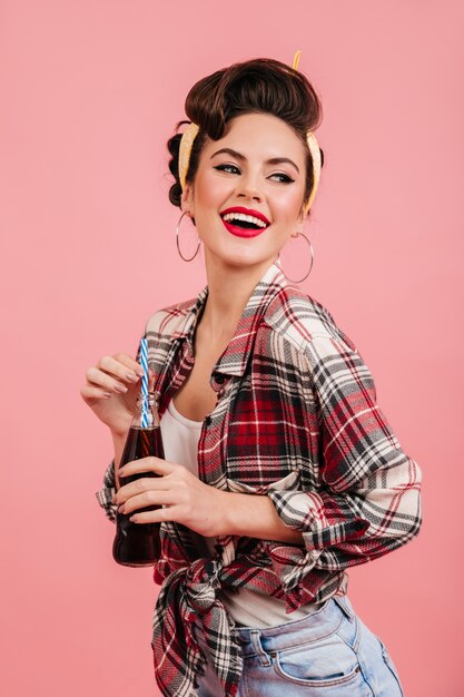 Adorable chica pinup expresando felicidad. Increíble mujer morena bebiendo bebidas sobre fondo rosa.