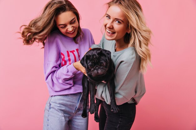 Adorable chica rubia posando con su hermana y bulldog francés. Sonrientes señoritas divirtiéndose con su mascota.