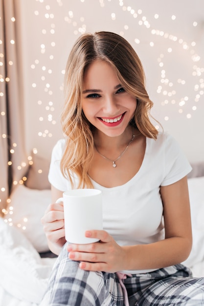 Adorable dama blanca en pijama riendo mientras bebe café. Filmación en interiores de increíble modelo femenino posando en el dormitorio con expresión de cara feliz.