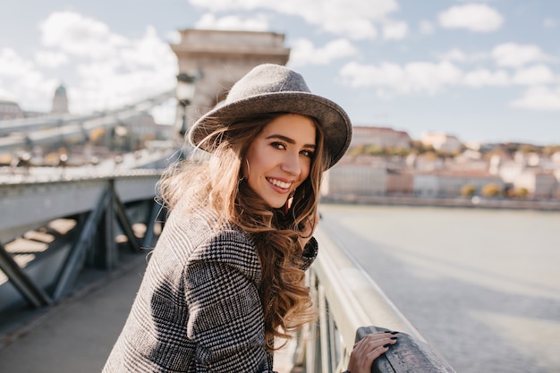 Adorable dama con cabello largo castaño claro posando en el puente sobre fondo borroso de la ciudad