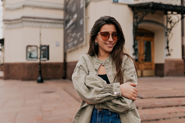 Adorable dama con cabello largo oscuro con chaqueta vaquera posando sobre edificios antiguos en el centro de la ciudad