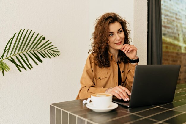 La adorable dama romántica con rizos con aspecto romántico está sentada en la mesa de la cafetería con una computadora portátil y una taza de café