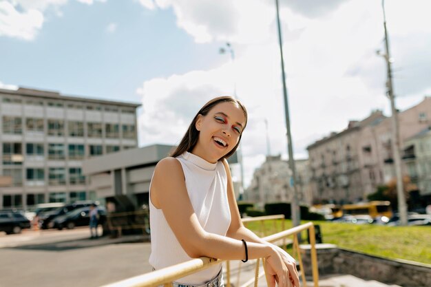 La adorable dama sonriente con cabello oscuro y maquillaje brillante está vestida con una camiseta blanca, sonríe y posa ante la cámara mientras camina por la calle