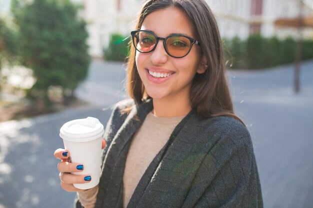 Adorable dama con una taza de café caminando afuera en un buen día soleado en la plaza de la ciudad