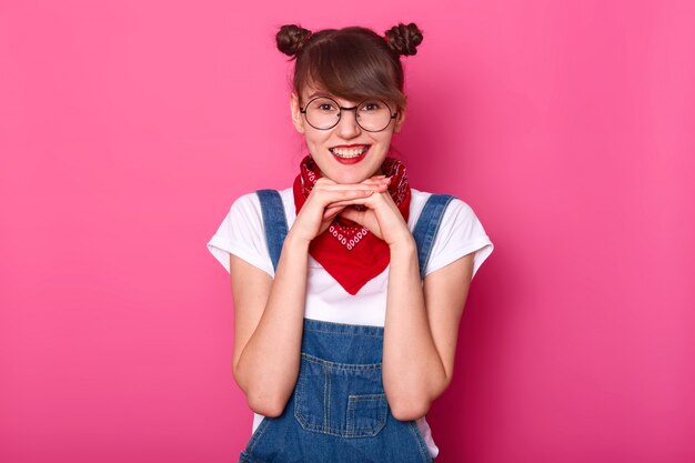 Adorable y encantadora joven estudiante tocando su barbilla con ambas manos, mirando directamente a la cámara, alegre, usando anteojos redondos y elegantes, overol de jeans, pañuelo rojo y camiseta blanca.