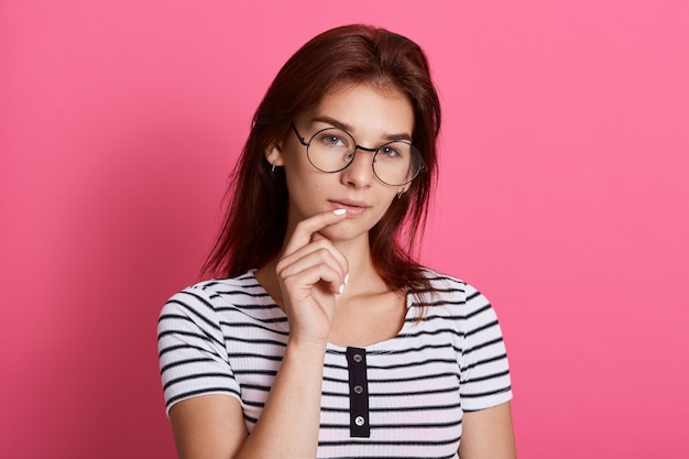 Adorable estudiante chica posando contra la pared color de rosa con expresión facial pensativa, vistiendo gafas y camiseta a rayas, manteniendo el dedo en los labios.