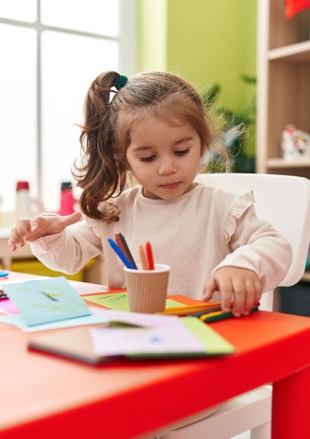 Adorable estudiante hispana sentada en la mesa dibujando en papel en el jardín de infantes